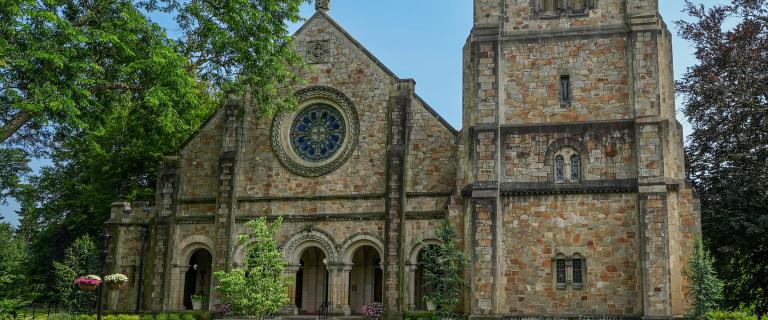 Exterior view of the Chapel at Vassar College featuring arched arcade entrances, a circular stained-glass rose window, and a prominent square bell tower flanked by lush green trees and blue sky.