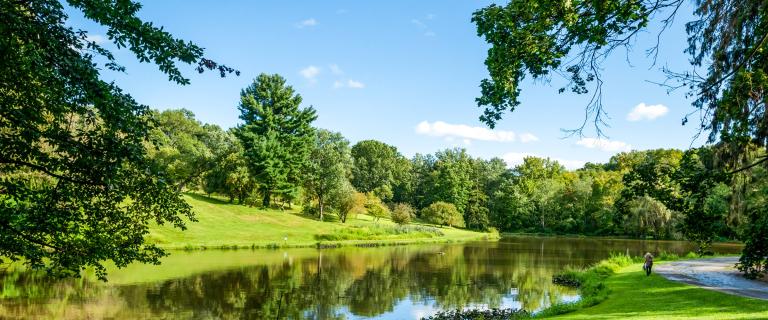 Scenic view of Sunset Lake at Vassar College bordered by grassy hills and lush green trees and a blue sky reflecting on the water.