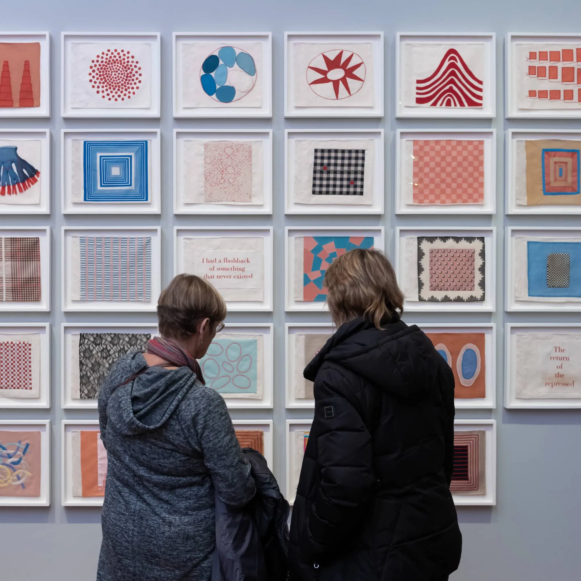 Two people, dressed in casual attire, are standing close to a wall, examining artwork consisting of multiple square frames, each containing different artistic patterns or designs, including abstract patterns, and varied color schemes.