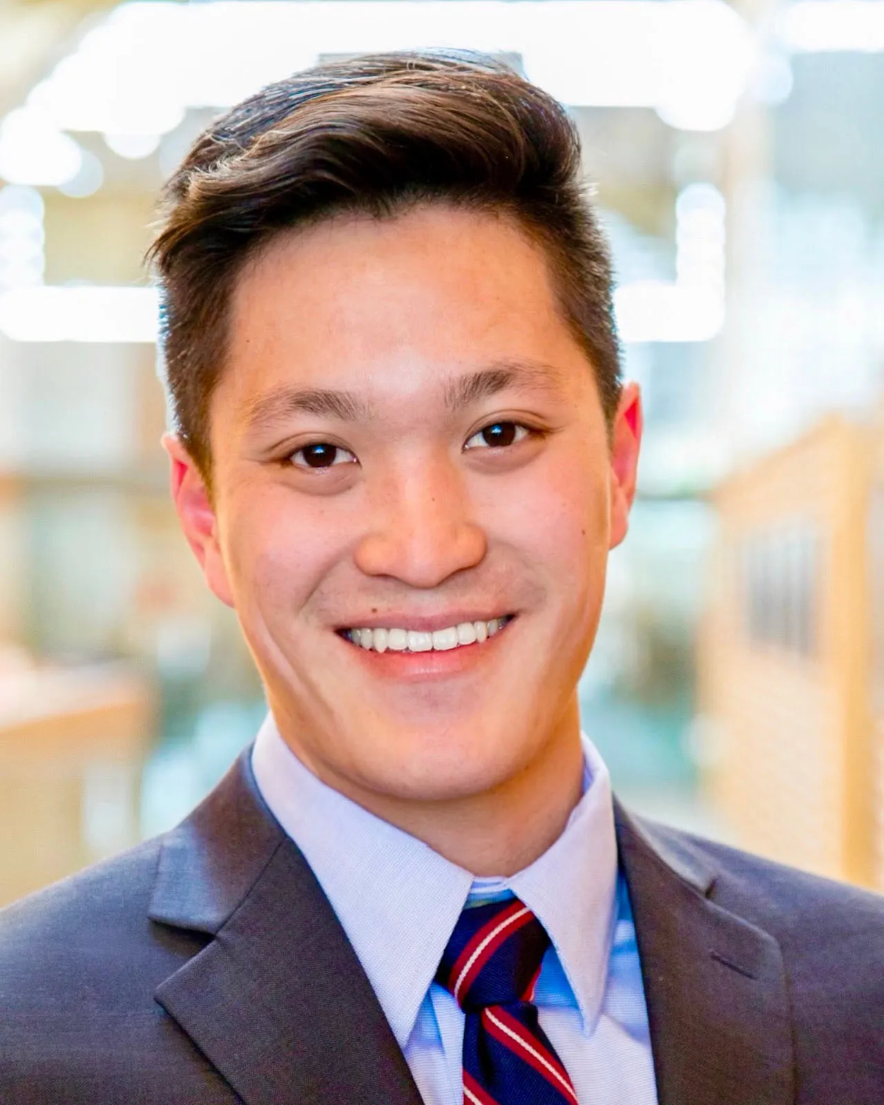 Kevin Lee ’14, a person with short, neatly styled dark hair wearing a suit jacket, light blue dress shirt, and red and blue striped tie, smiling in a brightly lit indoor setting with a blurred background.