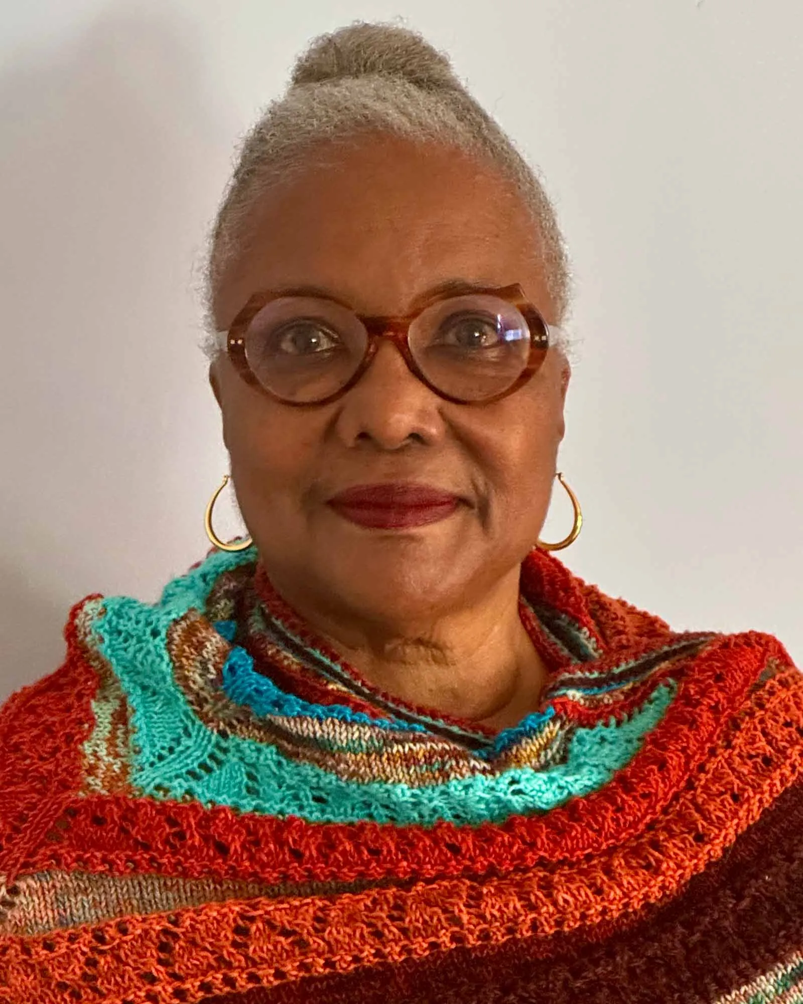 Maybelle Taylor Bennett ’70, a person with short, gray hair styled in a bun, wearing round eyeglasses, gold hoop earrings, and a multicolored knitted shawl in shades of red, orange, teal, and brown, standing against a plain light-colored background.