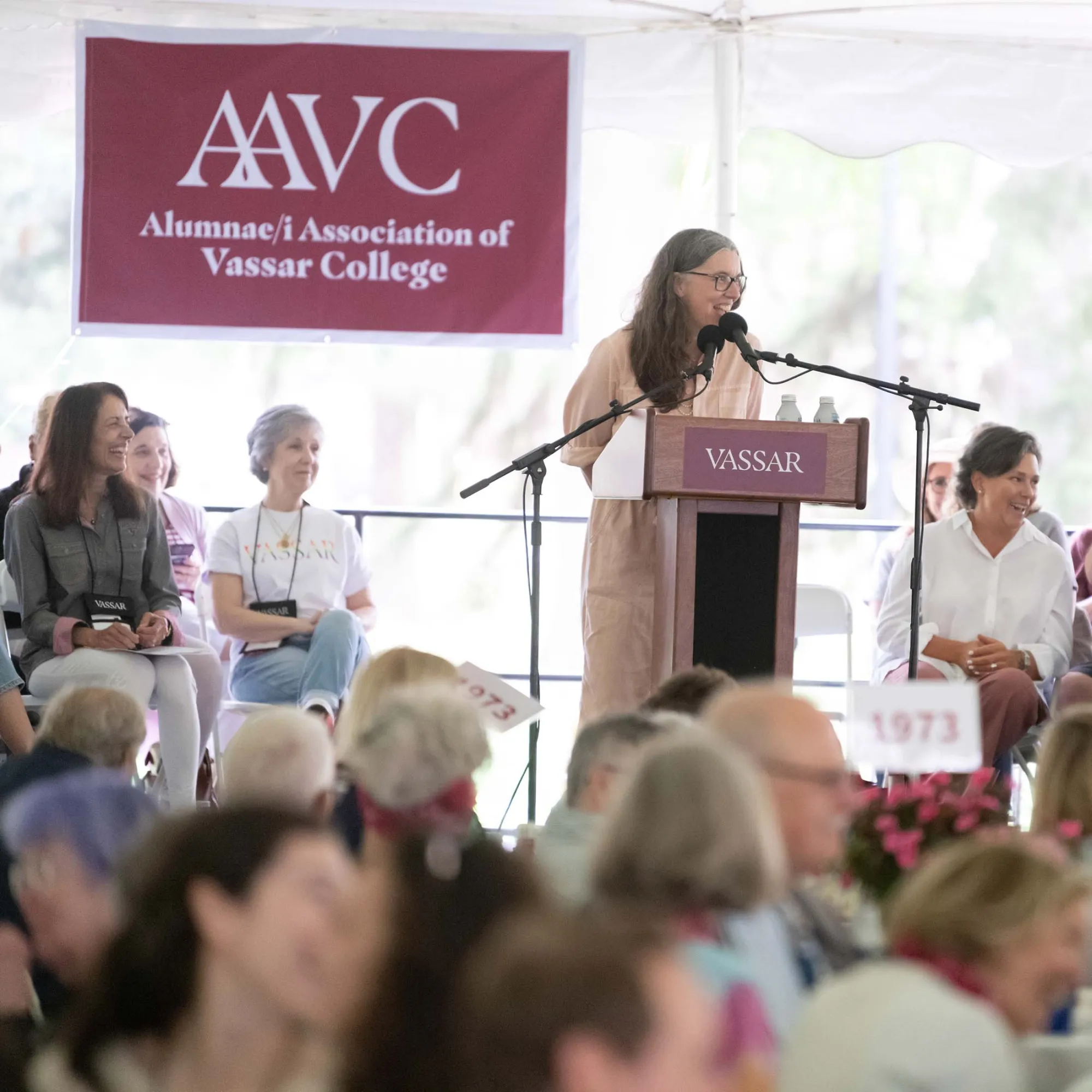 A person stands at a podium in front of a large group of people sitting under a tent, with a large banner behind them that says AAVC in white on a red background.