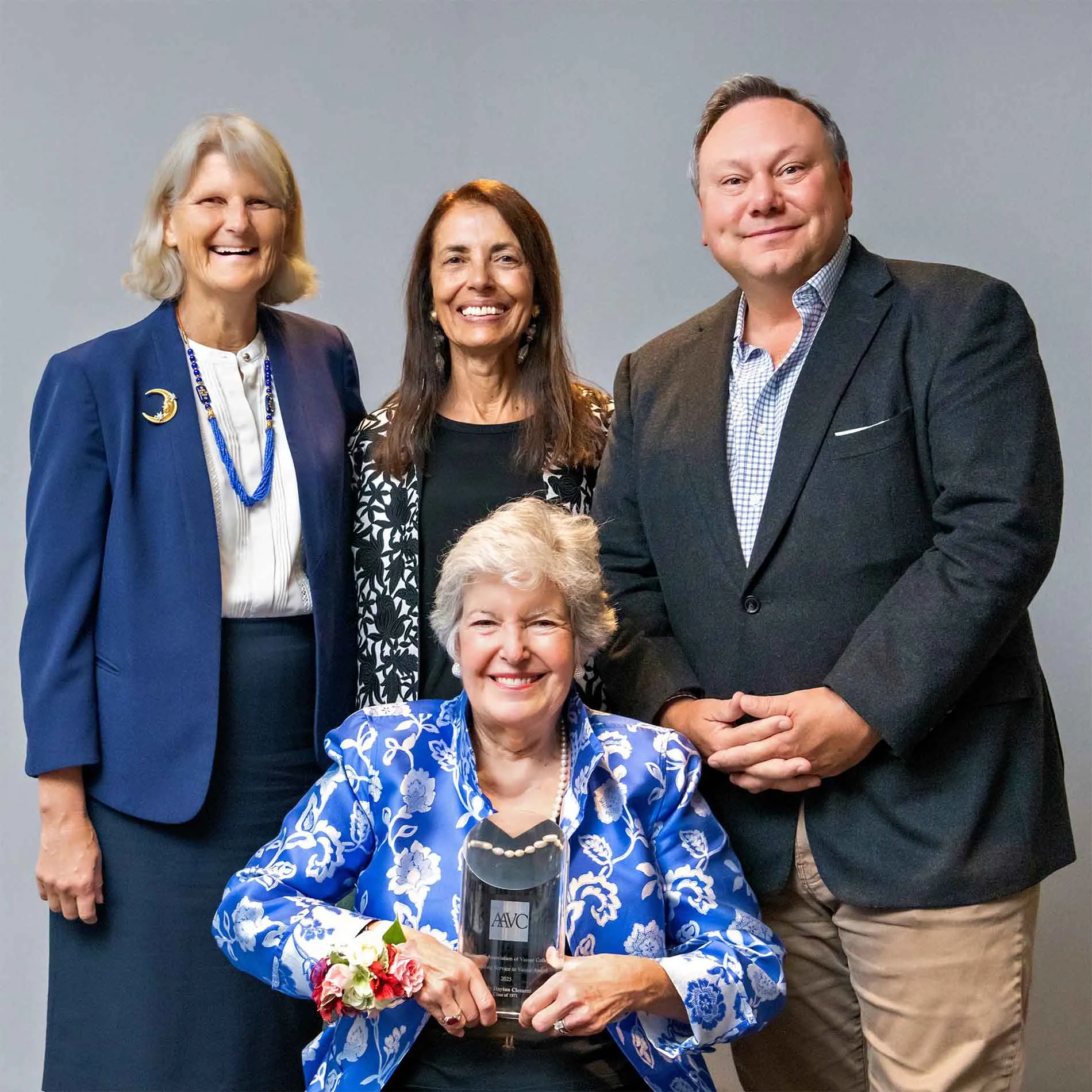 Four people posing together against a neutral background, one seated and holding an award, all smiling.