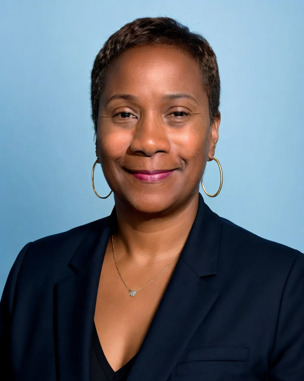 Professional headshot of a woman with short dark hair smiling at the camera. She is wearing a navy blazer over a black top, gold hoop earrings, and a delicate necklace. The background is a soft blue studio backdrop.