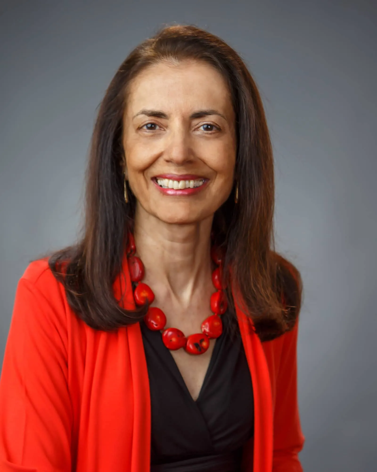 A person with long dark brown hair, a black shirt with a red sweater over it, and a red necklace smiles at the viewer.