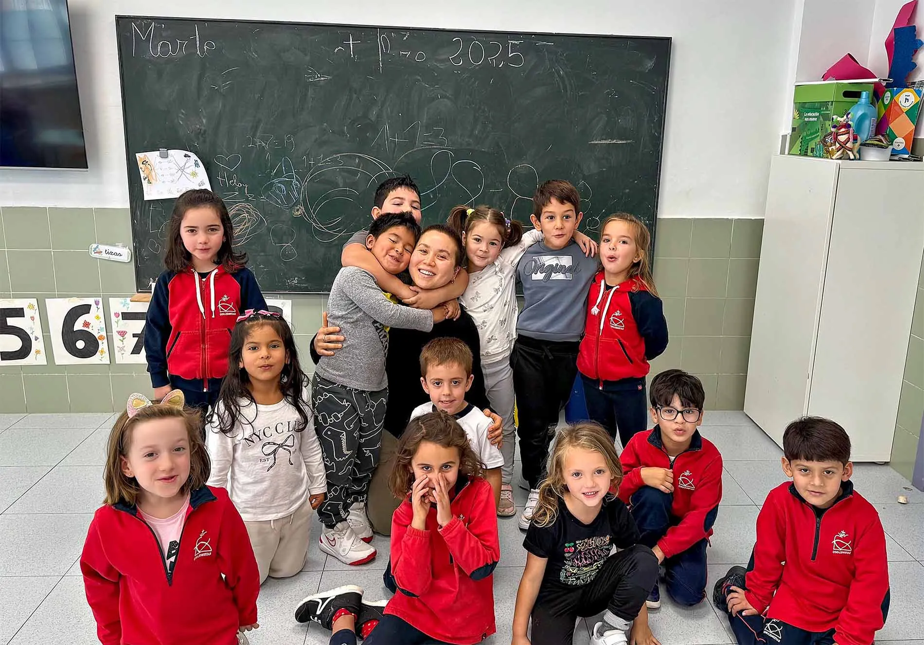 Teacher seated on the floor in a classroom, smiling while surrounded by a group of young children posing together in front of a chalkboard with drawings and writing.