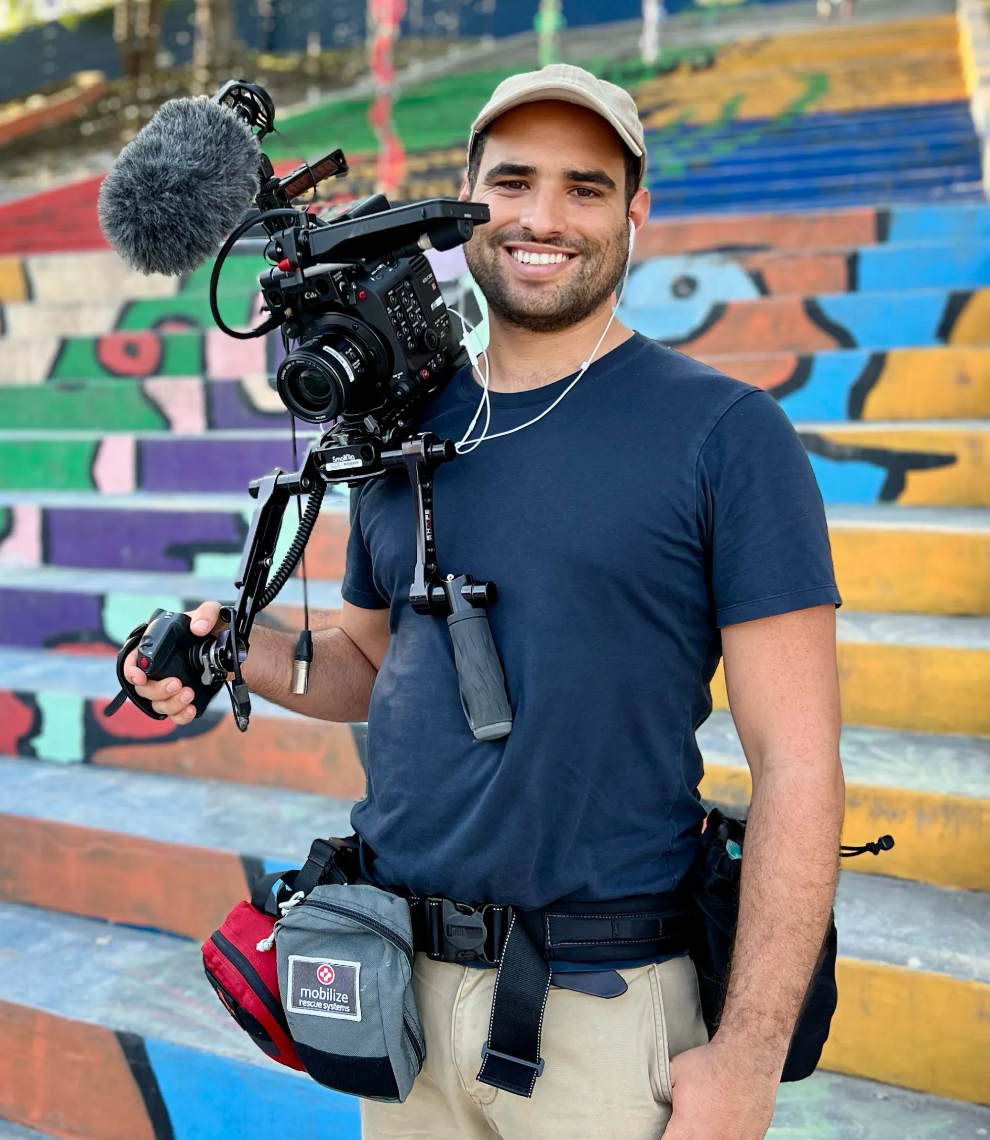A person wearing a cap, dark t-shirt, and belt with various pouches hanging from it smiles while holding a video camera rig equipped with a microphone and shoulder support, standing on brightly painted outdoor stairs.