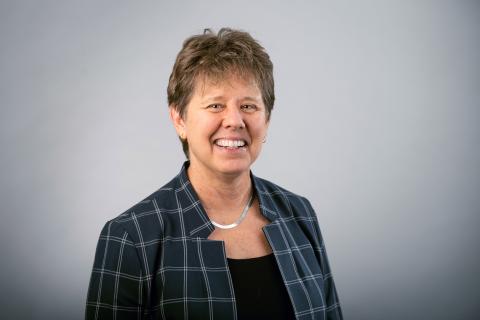 Jill S. Schneiderman wearing a black shirt, patterned dark blue shirt, and thick silver necklace against a light background.