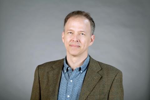 Mark Cleaveland wearing a light teal collared shirt and an olive colored jacket against a gray background.