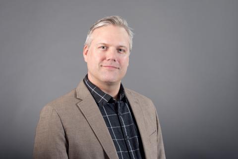 John Andrews wearing a black plaid shirt and beige jacket against a gray background.