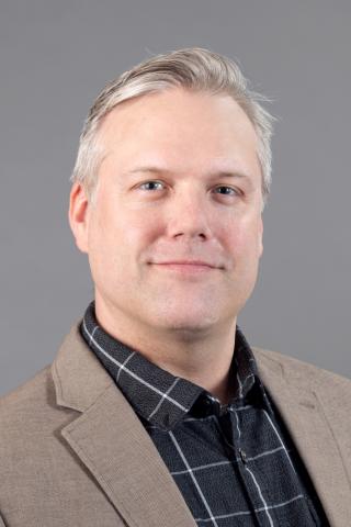 John Andrews wearing a black plaid shirt and beige jacket against a gray background.