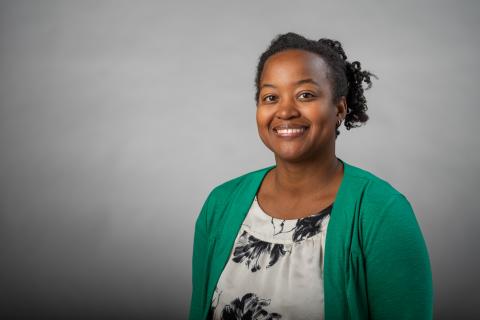 Kelli A. Duncan wearing a black and white patterned shirt and a green jacket with a gray background.