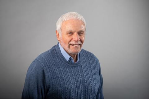 Sidney Plotkin wearing a light blue collared shirt and blue sweater against a gray background.