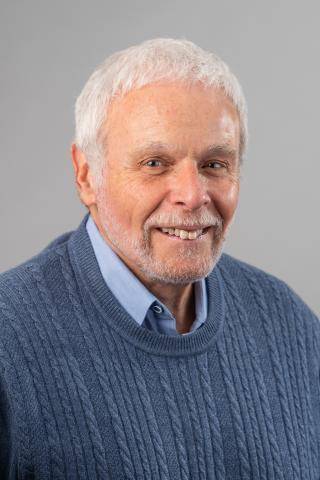 Sidney Plotkin wearing a light blue collared shirt and blue sweater against a gray background.