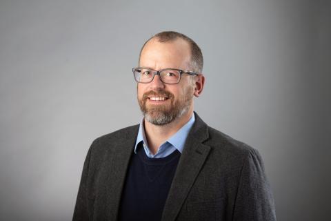 Zachary J. Donhauser wearing a light blue shirt, navy blue sweater and dark gray jacket against a gray background.