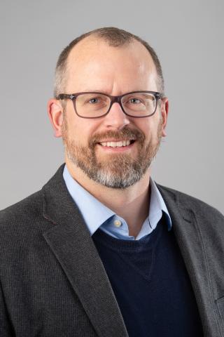 Zachary J. Donhauser wearing a light blue shirt, navy blue sweater and dark gray jacket against a gray background.