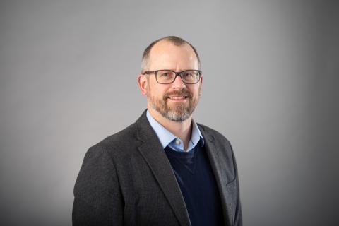 Zachary J. Donhauser wearing a light blue shirt, navy blue sweater and dark gray jacket against a gray background.