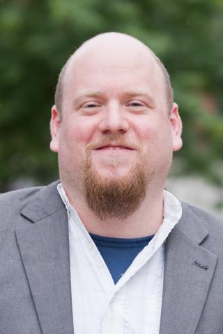 Alexander M. Kupfer wearing a white shirt and gray jacket with trees in the background.