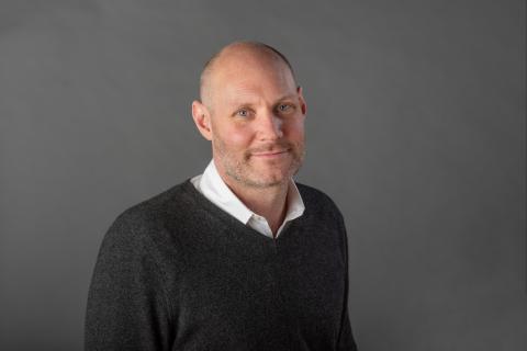 Hadley Creighton Bergstrom wearing a white collared shirt and dark gray sweater against a gray background.