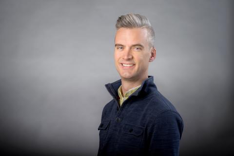 Colin Echeverría Aitken wearing navy shirt with high collar against a gray background.