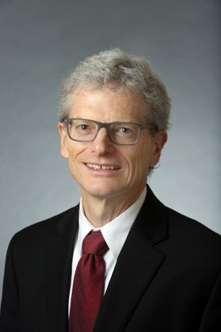 Jon Chenette wearing a white shirt, black jacket and red tie against a gray background.