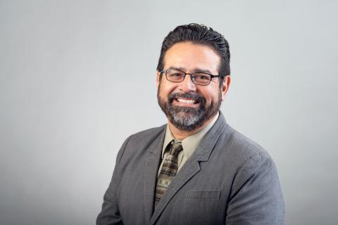 Jaime Liborio Del Razo wearing a light colored shirt, dark gray jacket against a gray background.