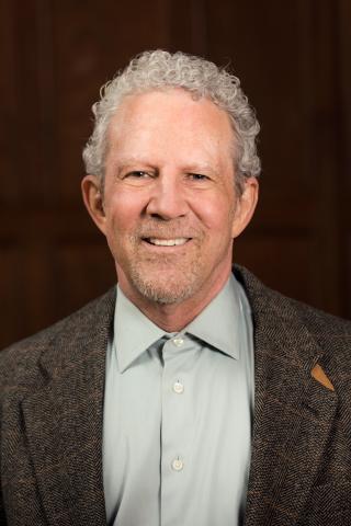 Brian J. Godfrey wearing a light colored shirt, dark gray jacket against a dark background.