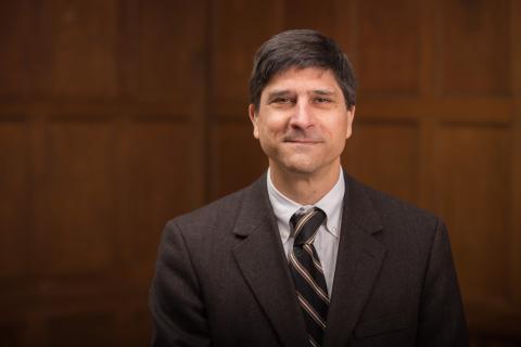 Ronald D. Patkus wearing a light colored shirt and brown jacket and tie with wood paneling in the background.