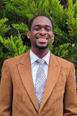 A person with short dark hair and facial hair smiles at the camera while wearing a tan suit coat and checkered tie.