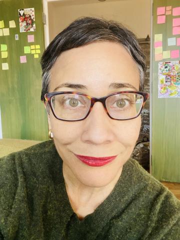 A close-up photo of a person with short dark hair, brown-rimmed glasses, and red lips smiling at the camera