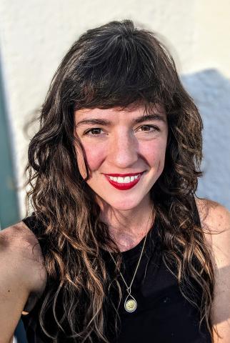 Woman with dark, curley hair, black dress, silver necklace, smiling for the camera.