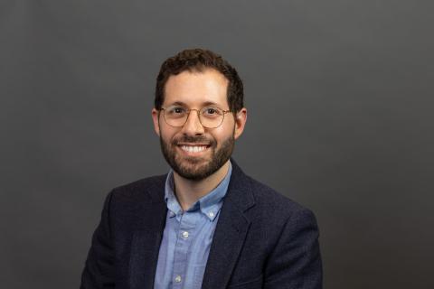 Headshot of person with dark curly short hair, glasses, and beard wearing a dark suit jack and light blue collared shirt.