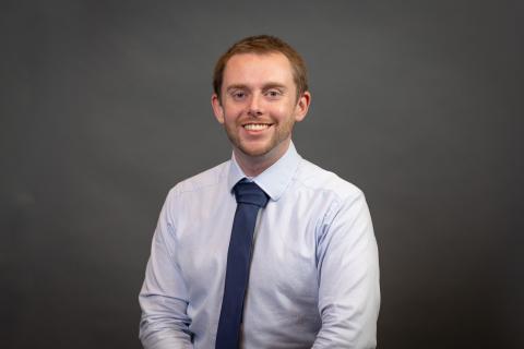 Person with short brown hair in a white collared shirt and blue tie smiling for a portrait,