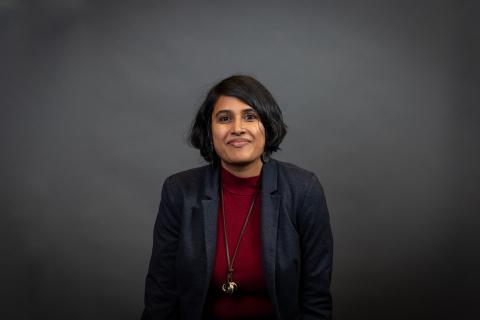 A person with medium length dark hair, dark skin, smiling, wearing a dark blue blazer, red mock-neck shirt, and long necklace with a gold pendant.