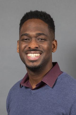 Deon Knights in a blue v-neck sweater smiling, against a light gray background..