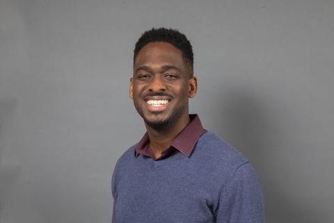 Deon Knights in a blue v-neck sweater smiling, against a light gray background..