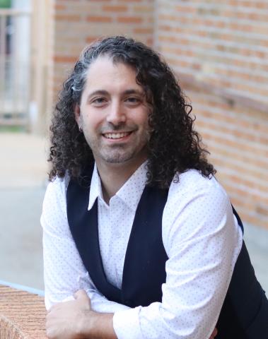 Person wearing white collared shirt and black vest with arms on a brick wall.