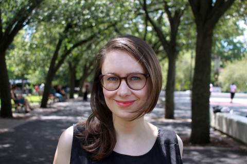 A woman with medium-length brown hair, wearing glasses and a black sleeveless shirt, smiling at the camera. The background shows a pathway lined in trees with green leaves.