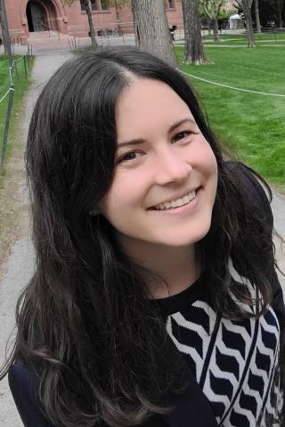 Woman with long black hair smiling outside with an outdoor path abd campus brick building in the background.