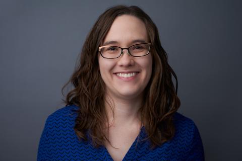 Person standing outside in the grass in front of a building wearing a light blue button shirt. The person is smiling with long brown hair and glasses.