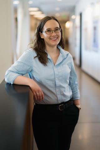 Person standing in a hallway with their arm on a shelf wearing a light blue button shirt. The person is smiling with long brown hair and glasses.