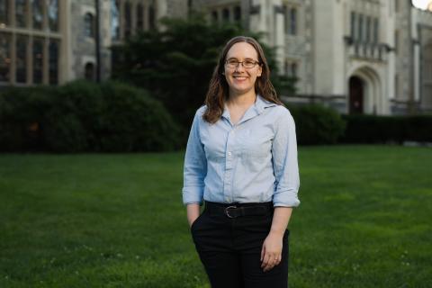 Person standing outside in the grass in front of a building wearing a light blue button shirt. The person is smiling with long brown hair and glasses.