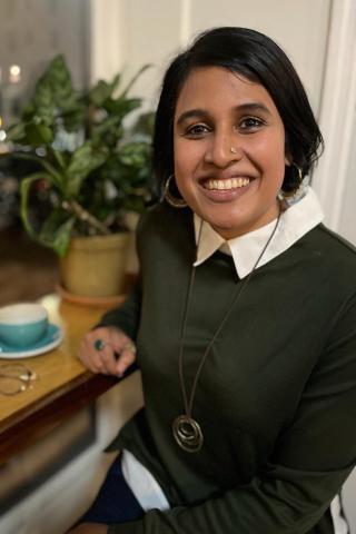 Arpitha Kodiveri wearing a white collared shirt under a green shirt, sitting with arm on a table with a blue tea cup and eyeglasses with a large green plant in the background.