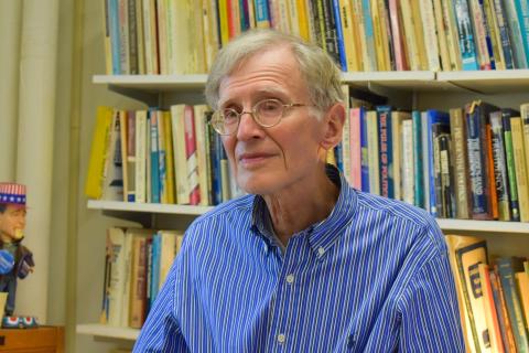 A person wearing glasses and a blue striped shirt sits in front of a bookshelf filled with books.