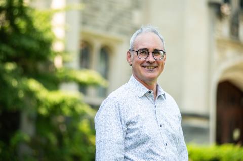 Portrait of a smiling individual in glasses, wearing a light patterned shirt, standing outdoors.