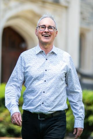 Portrait of a smiling individual in glasses, wearing a light patterned shirt, walking towards the camera outdoors.