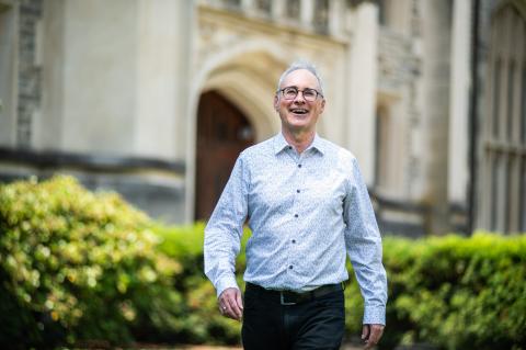 Portrait of a smiling individual in glasses, wearing a light patterned shirt, walking towards the camera outdoors.