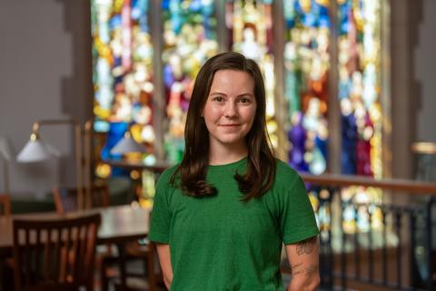 Person in a green shirt standing in the Vassar Library in front of a stained glass window.