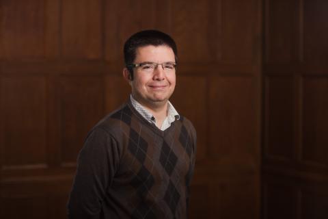 Portrait of a person with short dark hair and glasses, dressed in a brown argyle sweater over a white button-down shirt, standing against a paneled wood background.