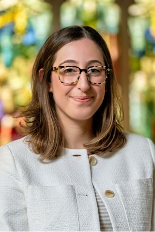 Stephanie Jackvony with shoulder-length brown hair and tortoiseshell glasses smiles while standing indoors, wearing a white textured jacket, with colorful stained-glass windows blurred in the background.